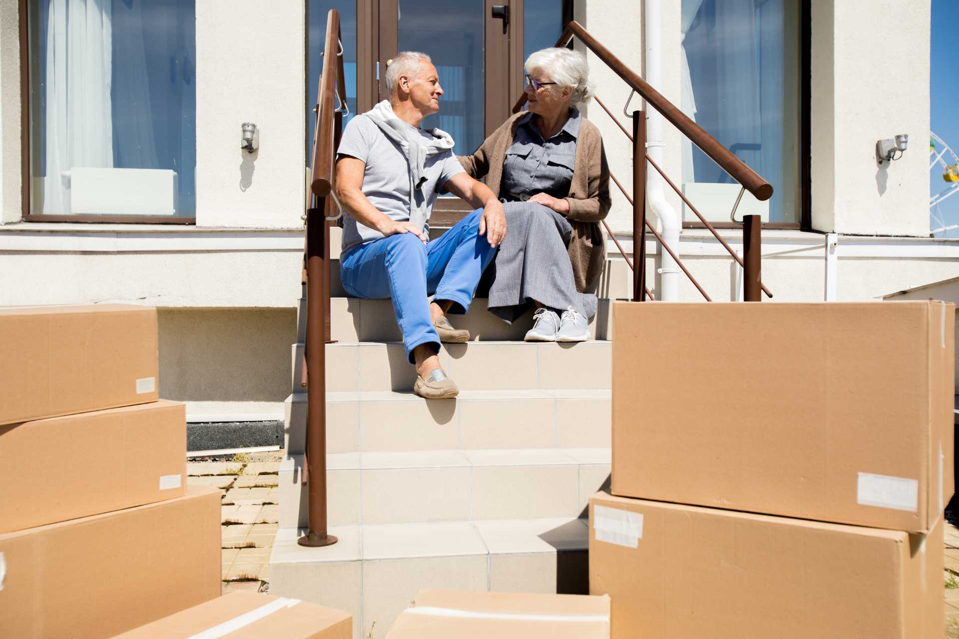Two smiling seniors sitting on their front stoop surrounded by moving boxes.